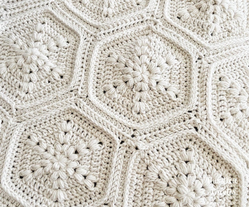 May include: A close-up of a crocheted blanket with a geometric pattern of beige hexagons. The blanket is made with a soft, textured yarn and has a subtle sheen.