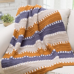 May include: A crocheted throw blanket with stripes of beige, orange, and purple. The blanket is made with a textured stitch and has a row of white bobbles along the bottom edge. The blanket is draped over a white couch. The text "This Pixie Creates" is visible in the bottom right corner of the image.