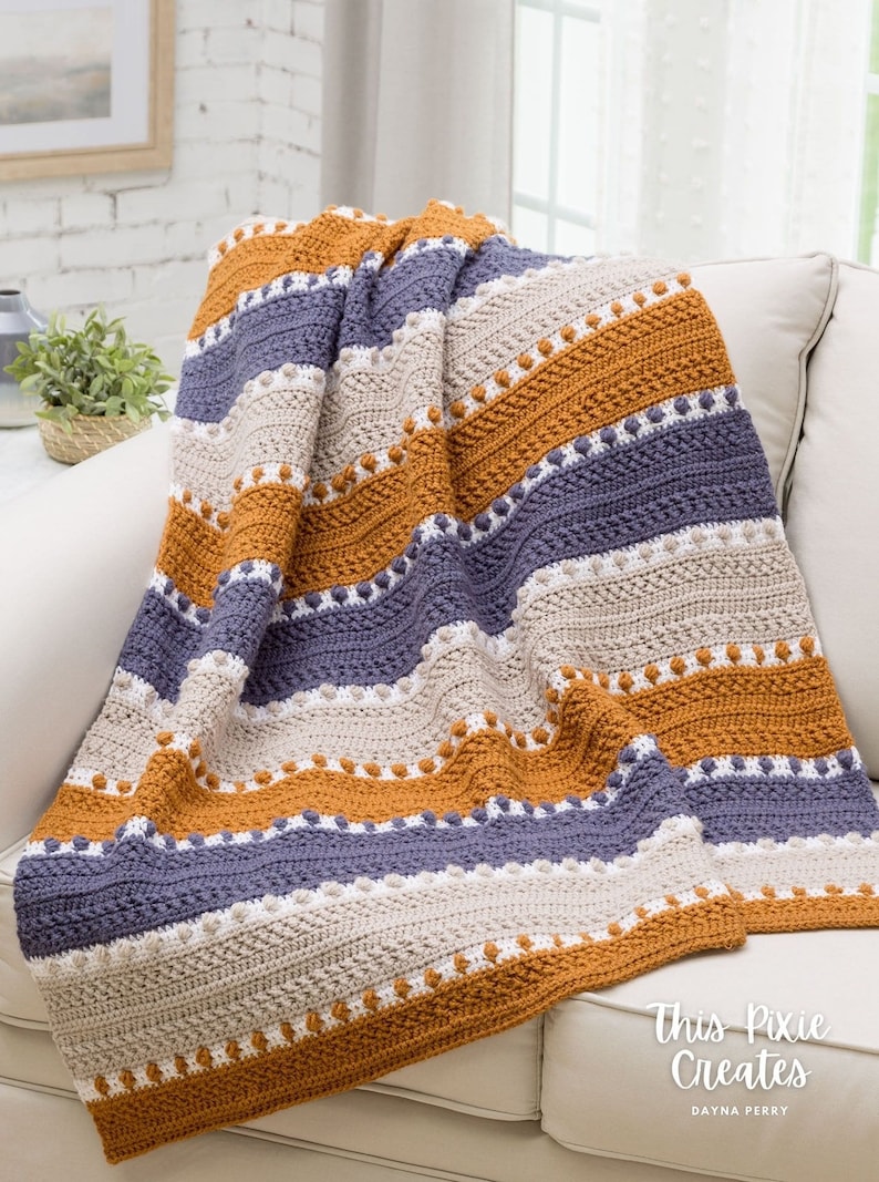 May include: A crocheted throw blanket with stripes of beige, orange, and purple. The blanket is made with a textured stitch and has a row of white bobbles along the bottom edge. The blanket is draped over a white couch. The text "This Pixie Creates" is visible in the bottom right corner of the image.
