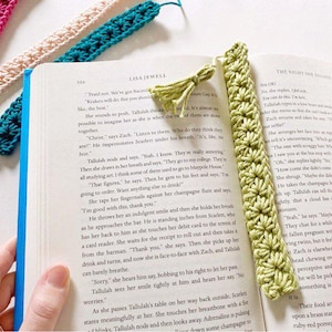 May include: A close-up of a green crocheted bookmark with a tassel. The bookmark is lying on an open book with text. The bookmark is made of a soft, textured yarn and has a delicate, intricate design.