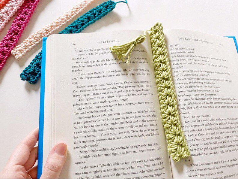 May include: A close-up of a green crocheted bookmark with a tassel. The bookmark is lying on an open book with text. The bookmark is made of a soft, textured yarn and has a delicate, intricate design.