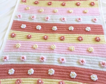 May include: A crocheted baby blanket with a striped pattern in pink, yellow, and white. The blanket is decorated with small white crocheted flowers.