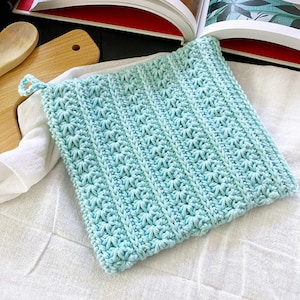 May include: A square, light blue crocheted potholder with a textured starburst pattern. The potholder has a loop for hanging and is resting on a white cloth. The handmade kitchen item is designed for heat resistance.