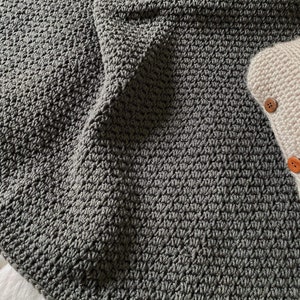 以下が含まれることがあります： テクスチャのあるパターンが施された、柔らかくグレーのクロシェ編みブランケット。ブランケットは半分に折り畳まれており、わずかに光沢があります。