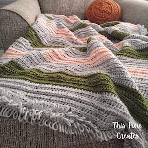 May include: A handmade crochet blanket with a striped pattern in gray, peach, and olive green. The blanket has a fringe trim and is draped over a gray sofa. The text "This Pixie Creates" is visible in the bottom right corner.