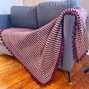 May include: A striped crocheted throw blanket with a burgundy and cream color scheme. The blanket has a fringe border with pom-poms.