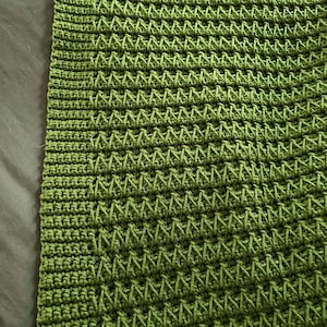 Op de afbeelding: Een groen gebreid deken met een textuurpatroon. Het deken is gemaakt met één kleur garen en heeft een gladde, gelijkmatige textur.