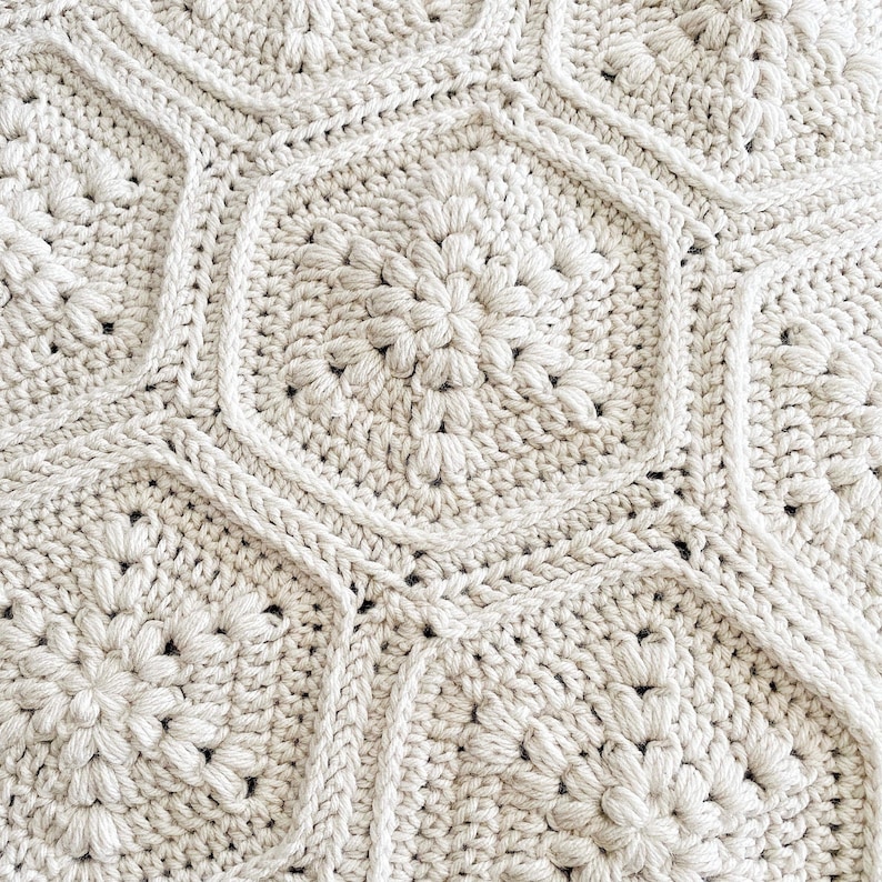 May include: A close-up of a crocheted blanket with a textured pattern of repeating flower shapes. The blanket is made of a cream-colored yarn.