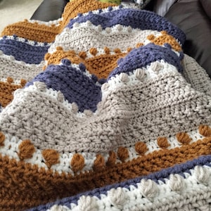 May include: A crocheted blanket with stripes of brown, gray, white, and blue. The blanket has a textured pattern with raised bumps and is folded on a couch.