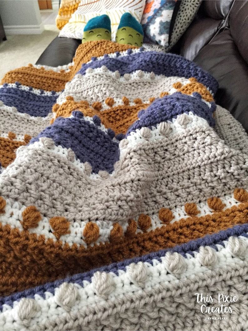 May include: A crocheted blanket with stripes of brown, gray, white, and blue. The blanket has a textured pattern with raised bumps and is folded on a couch.