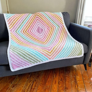 Puede incluir: Una manta de ganchillo colorida con un patrón de diamante en tonos de rosa, naranja, amarillo, verde y azul. La manta está colocada sobre un sofá gris.