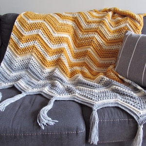 May include: A crocheted throw blanket with a chevron pattern in shades of mustard yellow, white, and gray. The blanket has fringe on the edges and is draped over a gray couch.