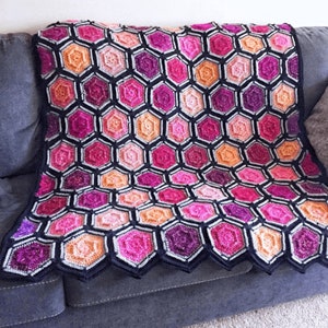 May include: A crocheted blanket with a honeycomb pattern in shades of pink, orange, and purple. The blanket is made with black yarn and has a scalloped edge.
