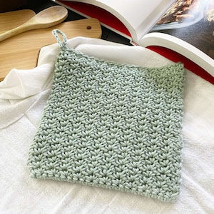 May include: A square, light green crocheted potholder with a loop for hanging. The potholder has a textured stitch pattern and is displayed on a white cloth. A wooden spoon and cutting board are in the background.