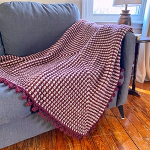 May include: A crocheted throw blanket with a burgundy and cream color pattern. The blanket has a fringe border with tassels. The blanket is draped over a gray couch.
