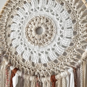 Puede incluir: Un atrapasueños de crochet blanco y beige con un aro de madera y borlas marrones, blancas y beige.