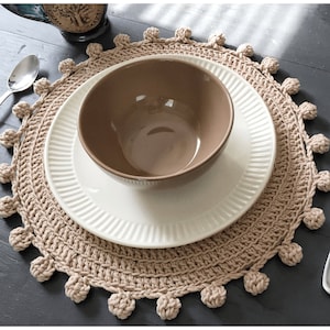 Puede incluir: Un mantel individual de crochet beige con un plato blanco y un tazón marrón en la parte superior. El mantel individual tiene un fleco de pompones alrededor del borde.