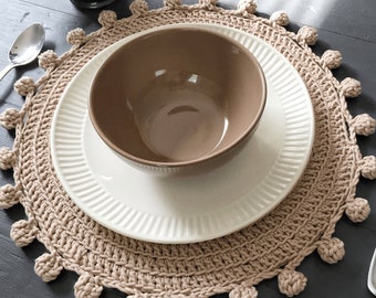 May include: A beige crocheted placemat with a white plate and a brown bowl on top. The placemat has a fringe of pom-poms around the edge.