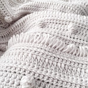 May include: Close-up of a white crocheted blanket with a textured pattern and a fringe border.