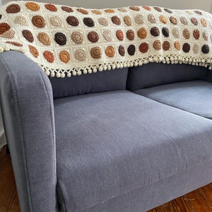 K&ouml;nnte beinhalten: Eine geh&auml;kelte Wolldecke mit Fransenbord&uuml;re. Die Decke ist aus beige, braunem und orangefarbenem Garn gefertigt und zeigt ein sich wiederholendes Muster aus Kreisen. Die Decke liegt &uuml;ber einem grauen Sofa.