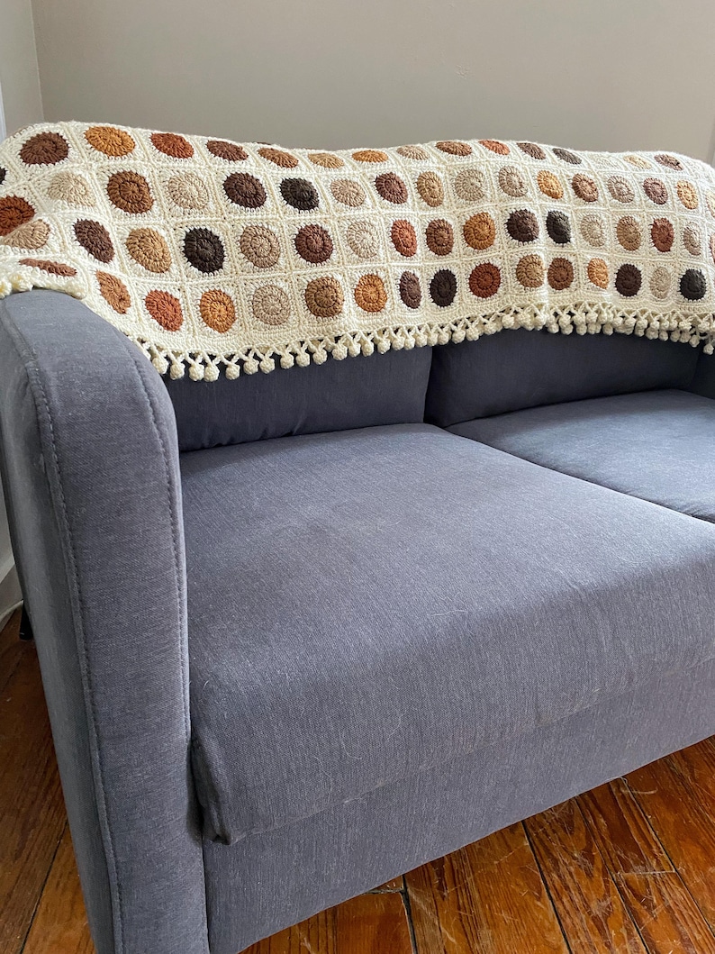 K&ouml;nnte beinhalten: Eine geh&auml;kelte Wolldecke mit Fransenbord&uuml;re. Die Decke ist aus beige, braunem und orangefarbenem Garn gefertigt und zeigt ein sich wiederholendes Muster aus Kreisen. Die Decke liegt &uuml;ber einem grauen Sofa.