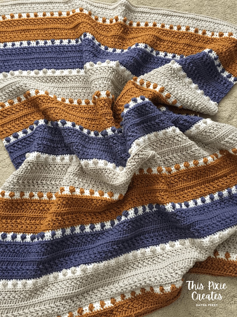 May include: A crocheted blanket with stripes of beige, brown, blue, and white. The blanket has a textured pattern with small bobbles.