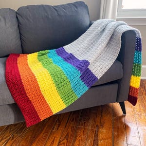 May include: A rainbow striped crochet blanket draped over a gray couch. The blanket is made with different shades of red, orange, yellow, green, blue, purple, and gray yarn.