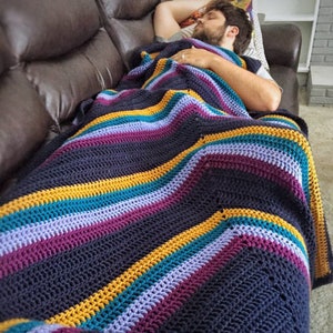 May include: A striped crocheted blanket in shades of navy blue, purple, teal and gold. The blanket is draped over a brown leather sofa.