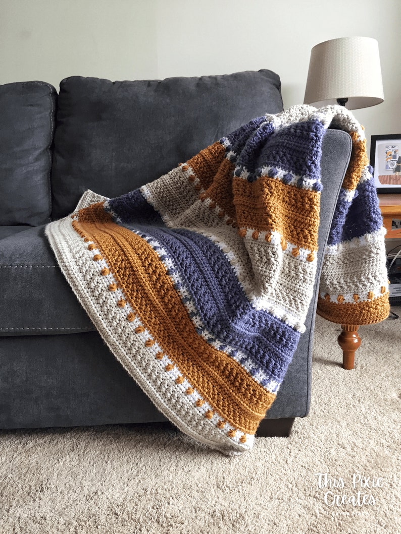 May include: A striped crocheted throw blanket in shades of beige, mustard yellow, and navy blue. The blanket is draped over a gray couch.