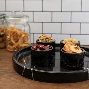 May include: A black marble serving tray with four small black marble bowls filled with dried fruit, nuts, and cranberries. The tray is on a wooden surface.