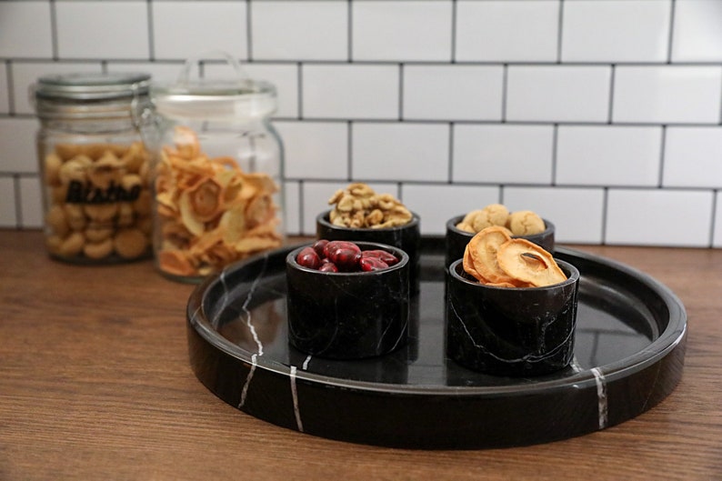 May include: A black marble serving tray with four small black marble bowls filled with dried fruit, nuts, and cranberries. The tray is on a wooden surface.