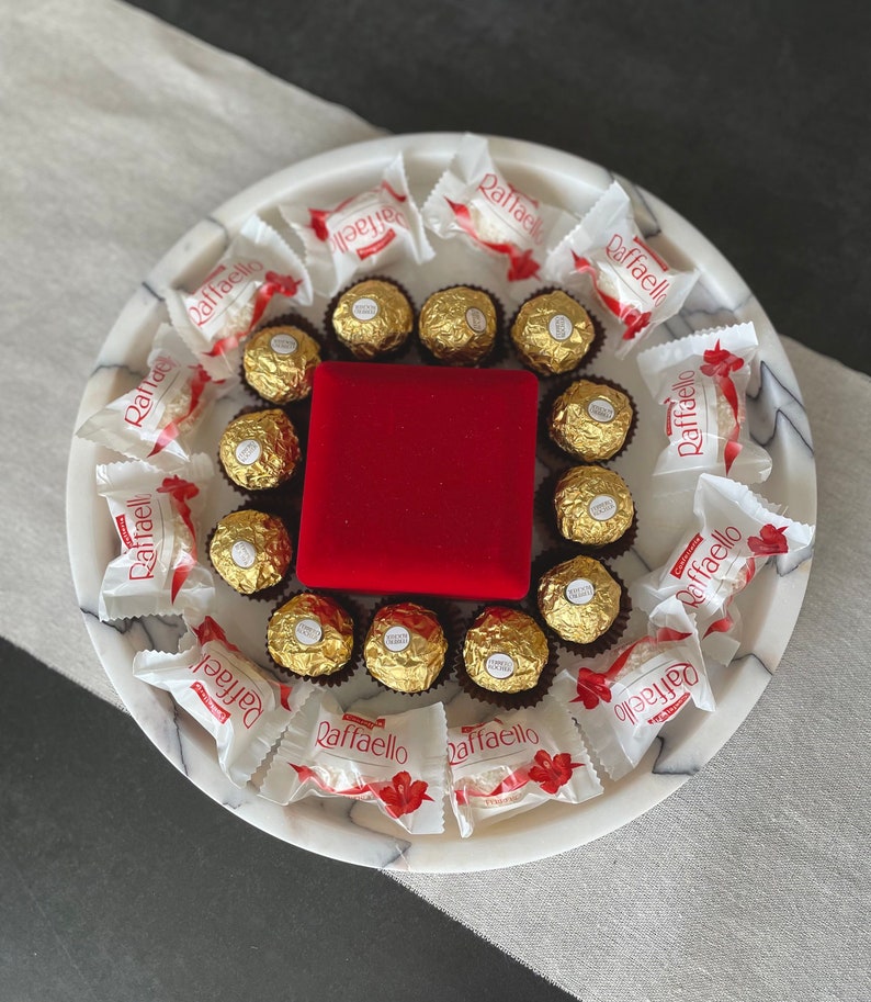 May include: A white marble serving tray with a red velvet box in the center, surrounded by individually wrapped chocolate candies. The candies are wrapped in white and red foil with the word "Raffaello" printed on them.