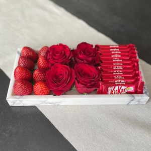Peut inclure: Un plateau en marbre blanc rempli de roses rouges, de fraises et de barres chocolatées Kit Kat. Le plateau est posé sur une surface blanche et grise.