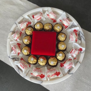 May include: A white marble serving tray with a red velvet box in the center, surrounded by individually wrapped chocolate candies. The candies are wrapped in white and red foil with the word "Raffaello" printed on them.