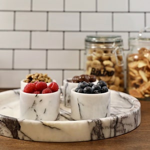May include: A white marble serving tray with four small white marble bowls filled with raspberries, blueberries, walnuts, and chocolate candies. The tray is on a wooden surface.