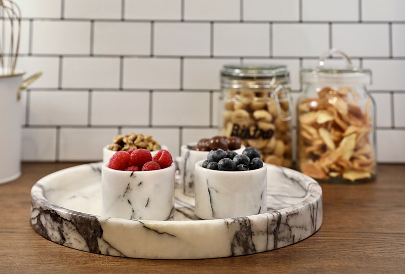 May include: A white marble serving tray with four small white marble bowls filled with raspberries, blueberries, walnuts, and chocolate candies. The tray is on a wooden surface.