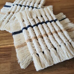 May include: Three handmade crocheted coasters with a fringe border. The coasters are made with natural white yarn and feature stripes of dark blue and light brown yarn.