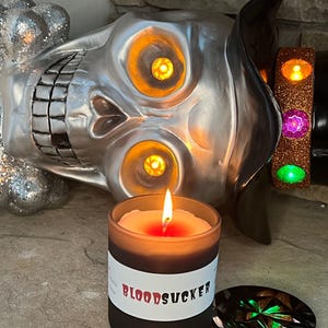 May include: A black candle with a white label that reads "BLOODSUCKER" and a lit wick. The candle is sitting on a table next to a silver skull with glowing orange eyes and a black hat with a green and purple jeweled band.