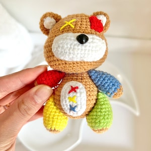 May include: A handmade crochet bear with brown, white, red, yellow, blue, and green accents. The bear has a white muzzle, black nose, and a yellow cross stitched on its head. The bear's limbs are different colors.