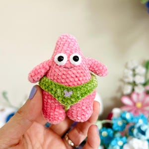 May include: A handmade pink crochet starfish figure with black eyes and a smiling face. It wears a green bikini with a purple heart. The plush toy is a whimsical, soft sculpture.