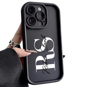 Peut inclure: Coque de téléphone noire avec une découpe pour l'appareil photo et les lettres "RS" en blanc, avec le nom "Reham Lamb" en écriture cursive. L'étui est tenu dans une main.