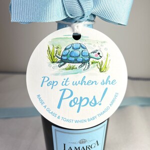 May include: A round white tag with a blue and green watercolor illustration of a turtle swimming in the water. The tag is tied to a bottle of sparkling wine with a blue ribbon. The tag reads "Pop it when she Pops! Raise a glass & toast when baby Thiago arrives."