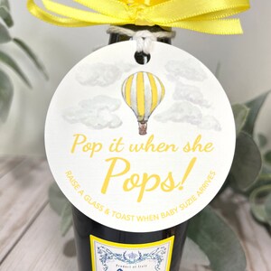 Puede incluir: Una etiqueta redonda blanca con una ilustraci&oacute;n de un globo aerost&aacute;tico a rayas amarillas y blancas. La etiqueta tiene el texto "Pop it when she Pops! Raise a glass & toast when baby Susie arrives."