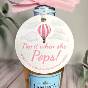 Puede incluir: Una etiqueta redonda blanca con una ilustraci&oacute;n de un globo aerost&aacute;tico rosa y blanco. La etiqueta tiene el texto "Pop it when she Pops! Raise a glass & toast when baby Olivia arrives".
