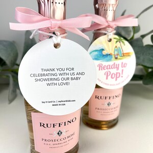 Może przedstawiać: Dwie małe butelki wina musującego r&oacute;żowego Ruffino Prosecco Rose z r&oacute;żowymi kokardkami i białą etykietą z napisem "Dziękujemy za świętowanie z nami i obdarowanie naszego dziecka miłością!" oraz białą etykietą ze sceną plażową i tekstem "Gotowy do strzelania!"