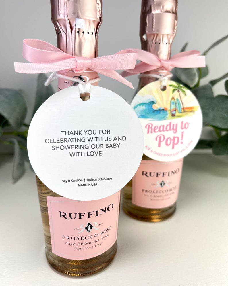 Może przedstawiać: Dwie małe butelki wina musującego r&oacute;żowego Ruffino Prosecco Rose z r&oacute;żowymi kokardkami i białą etykietą z napisem "Dziękujemy za świętowanie z nami i obdarowanie naszego dziecka miłością!" oraz białą etykietą ze sceną plażową i tekstem "Gotowy do strzelania!"