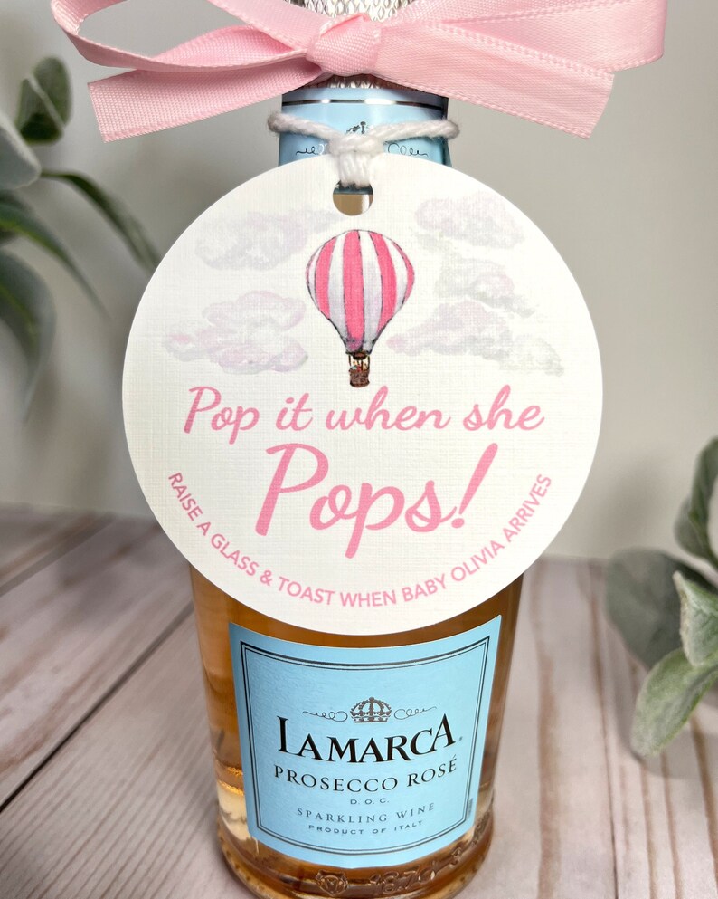 Puede incluir: Una etiqueta redonda blanca con una ilustraci&oacute;n de un globo aerost&aacute;tico rosa y blanco. La etiqueta tiene el texto "Pop it when she Pops! Raise a glass & toast when baby Olivia arrives".
