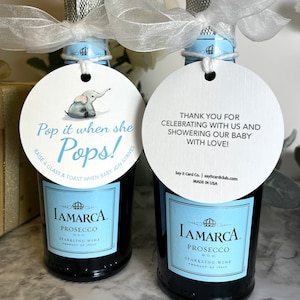 Two small bottles of prosecco with blue and white labels. Each bottle has a round white tag attached with a blue ribbon. The tag on the left bottle has a cartoon elephant and the text 'Pop it when she Pops! Raise a glass & toast when baby Jon arrives'. The tag on the right bottle has the text 'Thank you for celebrating with us and showering our baby with love! Pop It When She Pops Personalized Baby Shower Mini Champagne Bottle Favor Tag Prosecco Elephant Gift Tag Custom Baby Shower Gift Tag