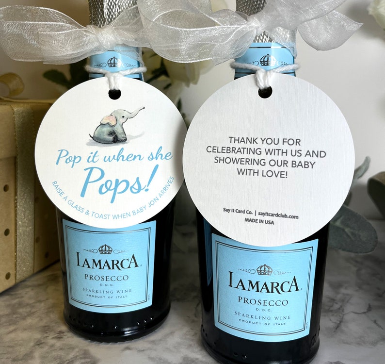 Two small bottles of prosecco with blue and white labels. Each bottle has a round white tag attached with a blue ribbon. The tag on the left bottle has a cartoon elephant and the text 'Pop it when she Pops! Raise a glass & toast when baby Jon arrives'. The tag on the right bottle has the text 'Thank you for celebrating with us and showering our baby with love! Pop It When She Pops Personalized Baby Shower Mini Champagne Bottle Favor Tag Prosecco Elephant Gift Tag Custom Baby Shower Gift Tag