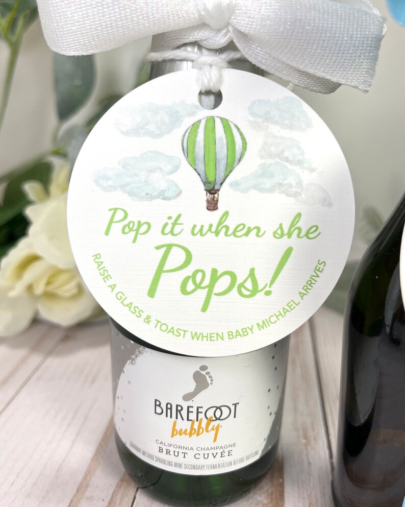 Puede incluir: Una etiqueta circular blanca con una ilustraci&oacute;n de un globo aerost&aacute;tico verde y blanco y el texto "Pop it when she Pops! Raise a glass & toast when baby Michael arrives".